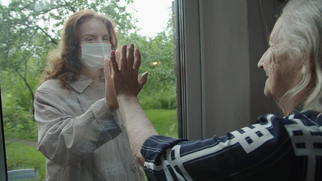 a woman in a protective mask visits her old mother in a country house with her hand to the glass to build safe contact and show support and trust during the covid-19 pandemic