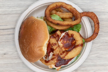 Overhead view of barbeque chicken sandwich stacked tall in the toasted bun with huge onion rings on the side to fill the entire plate