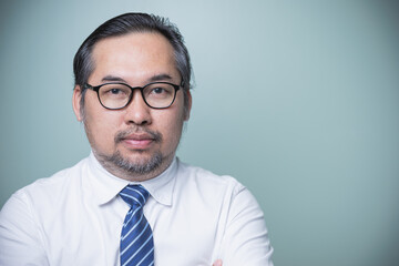 Portrait adult Asian businessman of high-confident wears a white shirt, tie blue, wearing glasses looking at camera while keeping arms crossed standing against on green background. Confident adult man