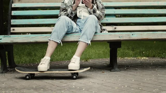 Beautiful, stylish girl-skateboarder sits on bench in city park while putting her legs on skateboard and using her cell phone
