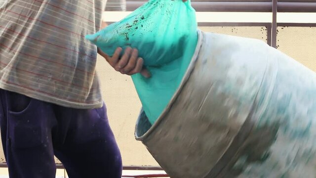 Detail of the arms and middle body part of a bricklayer dressed in casual clothes on the left of the frame, dumping a bag of sand into a cement mixer in operation located on the right of the frame