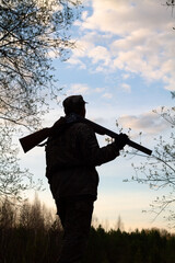a hunter with a discharged shotgun on his shoulder stands in the evening twilight