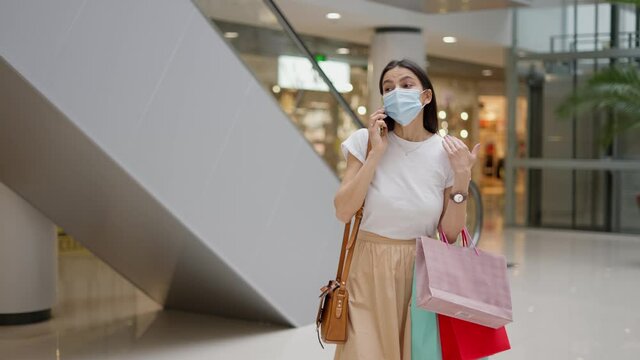 Beautiful caucasian female wearing protective face mask talking on modern smartphone with friend or family about successful shopping in mall after covid 19 quarantine, carrying many paper bag in hand