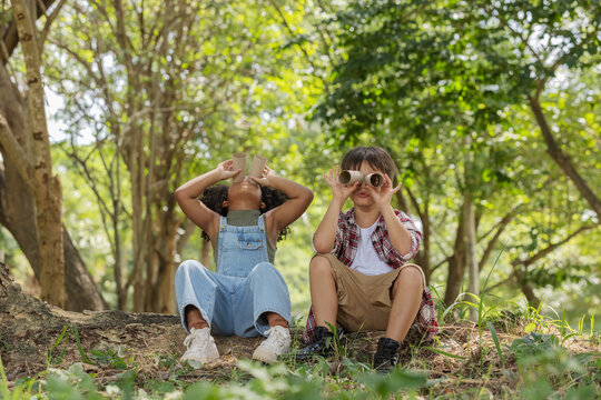 Multi-ethnic Children In Casual Clothing Sitting On Tree Roots, Looking At Through Tissue Tube Paper To Used Instead Binocular .Exploring Nature And Camping Summer Concept.