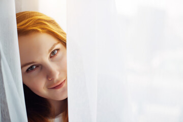 Cute young red-haired girl looks out from the white curtain, close-up, copy space. Portrait of beautiful smiling woman with perfect skin on a light blurry background.