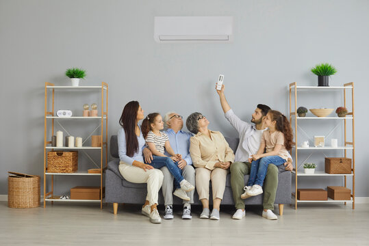 Happy Family Relaxing On Sofa Under Newly Installed Or Serviced Air Conditioner In Modern Home Interior. Young Parents, Kids, Senior Grandparents Adjusting AC And Enjoying New Cold Warm Mode Settings
