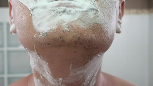 Unrecognizable male shaving foam on face using orange color razor. Men's beauty, skin care concept, white wall background. Facial hair removal in domestic bathroom. Care human skin