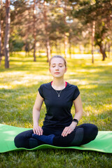 A young woman dressed in a sports uniform sits in a lotus position on a fitness mat in a city park.