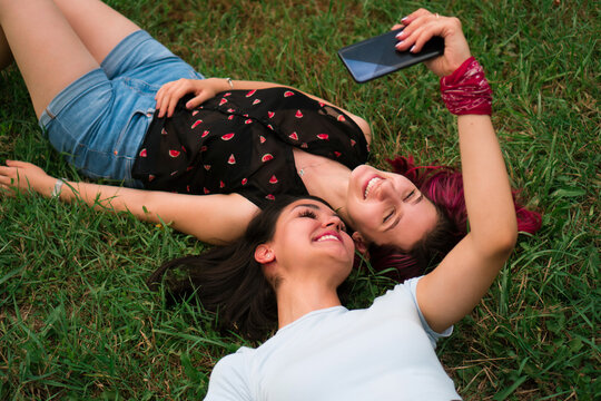 Two young girls are lying on the grass in the park and taking selfies with a mobile phone.