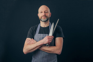 Bald male butcher or fishmonger in black t shirt and classic black and white apron on dark...