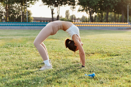 Portrait of slim young adult woman backbending on stadium field, working out outdoor, training alone, health care, healthy lifestyle, sport activity in summer.