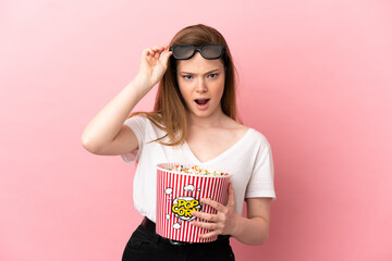 Teenager girl over isolated pink background surprised with 3d glasses and holding a big bucket of popcorns