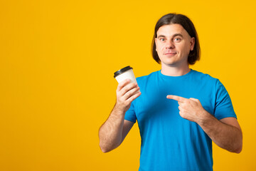 Portrait of young handsome caucasian man in blue shirt over yellow background holding cup of coffee to go. Banner copy space.