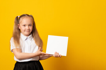 Education and school concept. Smiling little student girl with book. Copy space. Little kid looking at mockup poster and standing on yellow background.