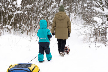 A woman with a boy in a turquoise overalls and a dog are walking in a snowy forest. People, lifestyle concept