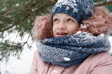 A teenage girl in a snow-covered blue hat and a pink jacket sits in the snow under a pine tree and smiles. People, lifestyle concept