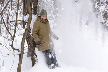 A woman in an olive-colored jacket and hat stands by the trees in a snowy forest under a snowfall and laughs. People, lifestyle concept