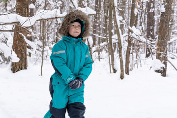 A boy in a turquoise overalls with a hood walks in a snowy forest and smiles. People, lifestyle concept