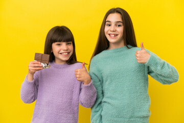 Little sisters isolated on yellow background taking a chocolate tablet and with thumb up