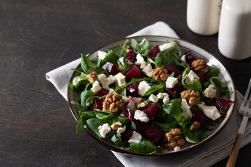 Beet or beetroot salad with fresh arugula, soft cheese and walnuts on plate, dressing and spices on dark wooden background, copy space, top view