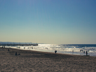 Fototapeta premium A beach filled with beach goers, playing on the sand and in the water, with a pier in the background and the sun reflecting off the water