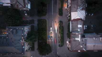 A white car with lights on drives along the city's evening street among houses and trees - an overhead drone shot.