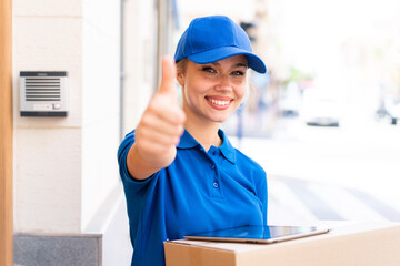 Young delivery woman at outdoors holding boxes and a tablet with thumb up
