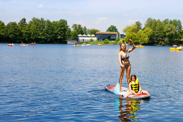 Fototapeta na wymiar Young woman with school girl paddling on sup board on a lake. Active family on modern trendy stand up paddle board. Summer outdoors vacations activity for family and children. Watersport activieties.