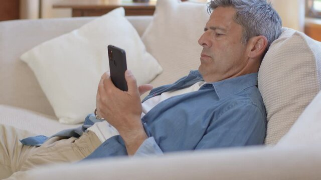 Medium Shot of Middle Aged Man Slouched On Sofa Using Smartphone 