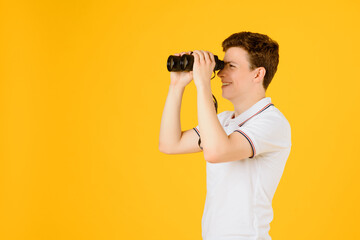 Portrait of a young happy man in white t-shirt smiling and looking through binoculars on a yellow background. Spyglass tourism concept. - obrazy, fototapety, plakaty
