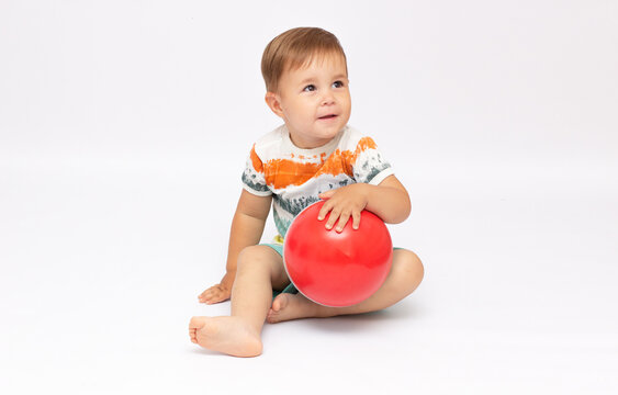 Playful cute little baby boy dressed in a striped bodysuit, sitting and playing a new ball on a white background. Concept of children's games. Place for text. Active childhood concept.