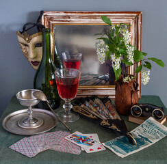 A romantic still life with a glass of red wine, an old theatrical advertisement, binoculars, an old mirror with a Venetian mask and a fan hanging on it. Vintage.