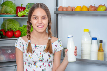 Beautiful young teen girl holding bottle of milk and drinks while standing near open fridge in kitchen at home