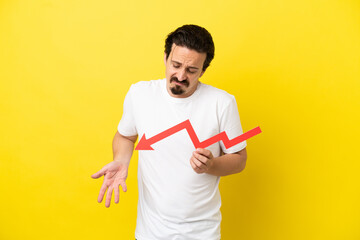 Young caucasian man isolated on yellow background holding a downward arrow and with sad expression