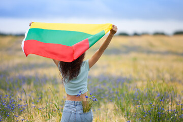 Young woman holding flag of Lithuania in a rye field with blue cornflowers. Back view. Lithuanian Flag Day. Independence restoration Day. Travel, learn lithuanian language and love Lithuania concept. - obrazy, fototapety, plakaty