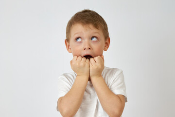 Child with horror looks up biting his hands in fear on a white background. Emotion of fear on the face of the boy with blue eyes