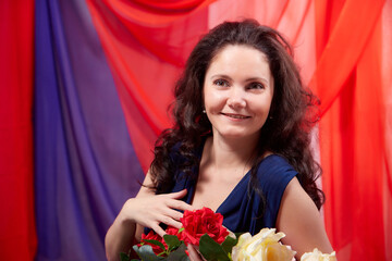 Woman in blue dress with long brunette curly hair and roses in hands indoors with red and blue background. Model posing in studio during photo shoot