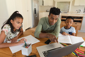 Happy african american father using laptop, daughter and son doing homework at home