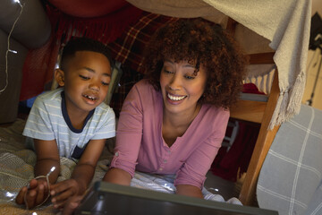 Happy african american mother with son at home, lying in blanket fort and using tablet