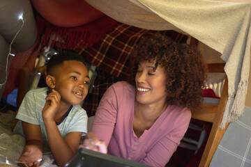 Happy african american mother with son at home, lying in blanket fort and using tablet