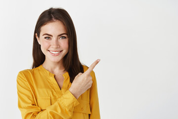 Close-up of brunette female model showing promo offer, pointing finger right and smiling at camera, white background - 442892959