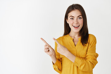 Excited young woman showing banner, pointing fingers left and smiling at camera, standing amazed at white background
