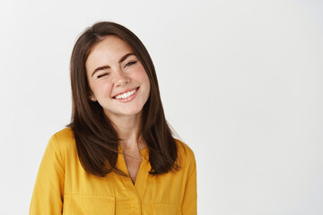 Close-up of happy young woman winking at camera, smiling joyful, standing over white background
