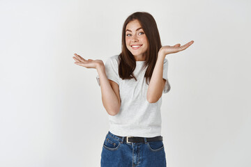 Smiling young woman shrugging shoulders and asking what, acting clueless and innocent, standing over white background