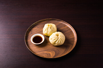 wooden plate with steamed Bao buns and bowl with soy sauce, over dark wooden table