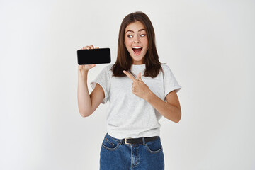 Excited smiling woman holding smartphone, showing blank cellphone screen and pointing at phone, standing over white background