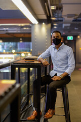 Fototapeta na wymiar Full length shot of of handsome black African businessman sitting inside shopping mall while wearing face mask to protect from covid-19