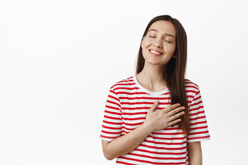 Obraz premium Image of dreamy smiling brunette girl, thinking of smth romantic, close eyes and hold hands on heart, remember, imaging smth beautiful, standing against white background
