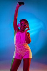 Young pretty African girl in sunglasses and headphones dancing isolated on blue studio background...