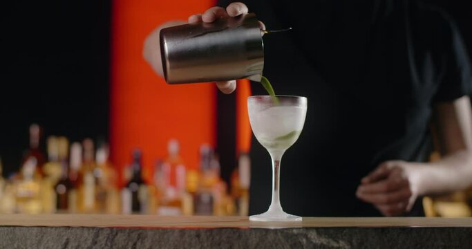 Bartender pours mixed drink from the shaker to the frozed high-stemmed glass, barman makes cocktail with ice and mixed alcohol, 4k 120 fps Prores HQ